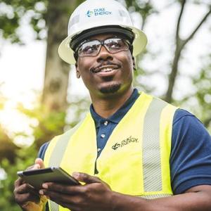 A person in safety hat and vest with a tablet in their hands, sun through the trees behind them.