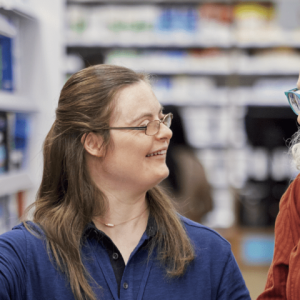 Two people smiling at each other in a pharmacy aisle, one points to an item on a shelf.