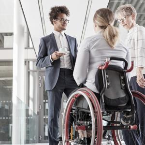 Two people standing and talking with a third in a wheelchair outside a glass-walled building.