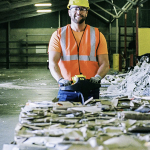 Worker in an orange safety vest and yellow helmet standing behind bales of paper recycling and smiling