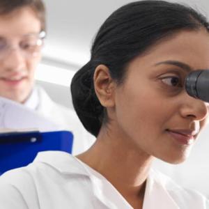 A person looking into a microscope, another looking on with a clipboard.