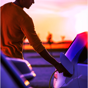 A person holding a plug to an electric vehicle port. A setting sun in the distance.