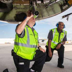 two people kneeling underneath an airplane, both wearing safety vests and hearing protection