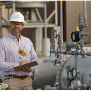 A person in a hard hat writing on a clipboard, next to industrial pipes outside.