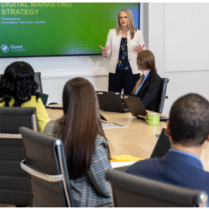 A person presenting to a room of others seated at a long table. A screen behind them.