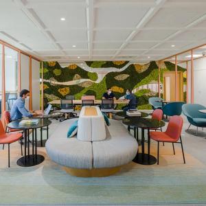 People working in an open space, comfortable chairs and bench seating, a mural of greens on one wall, lots of windows.