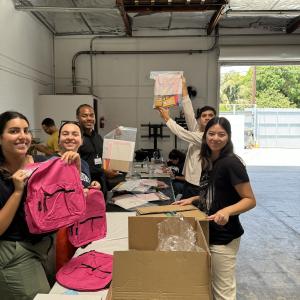 Volunteers standing around long tables holding boxes, pink backpacks, and other items for care packages