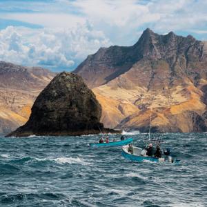 Two small boats carrying people on wavy water. Large mountains in the distance.