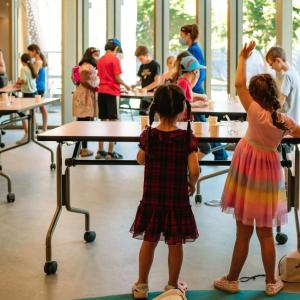Kids standing at tables, one with hand raised.