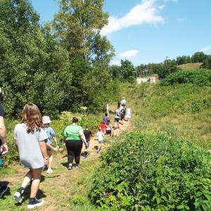 Kids and adults walk in a line along a narrow path in a lush area.