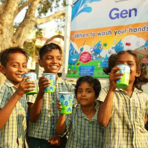 A group of smiling children holding out cups. A sign "Gen. When to wash your hands" behind them.