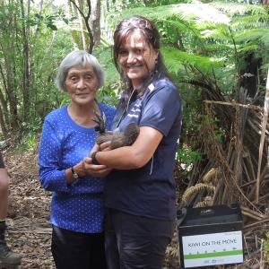 A person holding a Kiwi bird and another elder holding their arm. Both standing in a wooded area. A box with "Kiwi on the move" on the ground next to them.