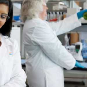 Two people in a laboratory setting, wearing white lab coats and protective gloves and eye wear. 