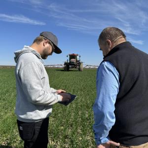 Two people standing together in a field looking at a screen, with a sprayer in the background