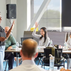 A person with hand raised speaking to a room of adults, some with their hands raised.