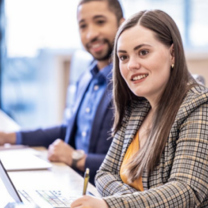 Two smiling people seated at a table, looking in the same direction, taking notes.