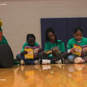 Children sitting in a line on a gym floor, a tree mascot next to them, all reading books.