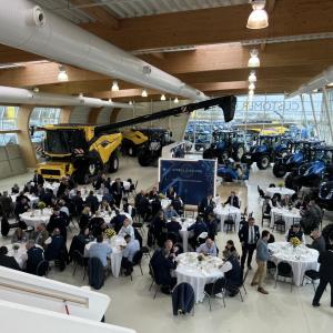 Wide view of a group of people seated at circular tables. Large farming and construction vehicles behind them.