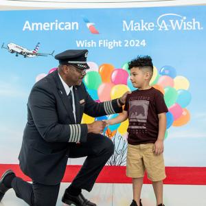 A pilot kneels and shakes the hand of a child. A poster behind them with American and Make a Wish logos, balloons.