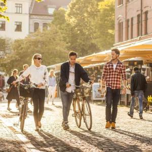 three people walk down a brick lined street, pedestrians and bike riders in the background on a sunny day