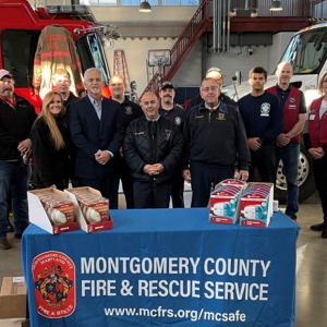 A line of people standing in front of a fire truck with a banner "Montgomery county fire and rescue service". A table with smoke detectors in front of them.