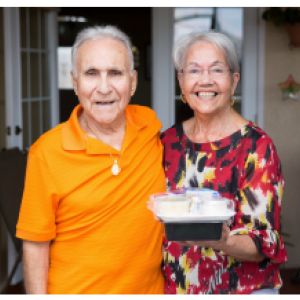 Two elderly people smiling on a porch, one holding a container of food.