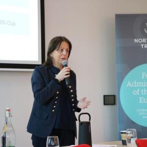 A speaker with a microphone, a large display on the wall behind them.