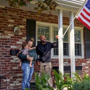 Two adults and a small child standing on a porch. An american flag in a post on the side pillar.