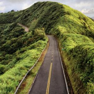 A two lane paved road leading through a greenery-covered peak. Cloudy sky in the background.