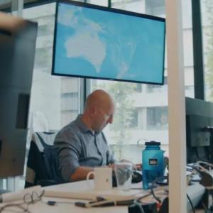 A person seated, working on a laptop in an office setting with many computers and monitors around.