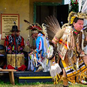 A person in ceremonial garments dancing, three behind them playing a drum.