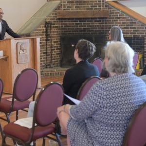 A person speaking at a podium in a room of others seated.