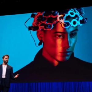A person speaking on a stage, a digital screen showing a person with device on their head.