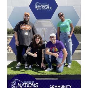Four people posing with a "Concacaf Nations League Finals, Comunity Day of Play" banner