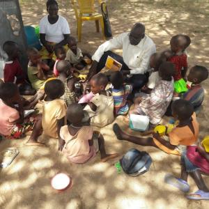 A group of children sitting on sandy ground, an adult showing them a picture