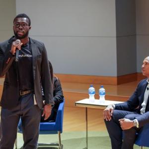 Two people seated on a stage, a third is standing in front of them with a microphone, speaking.