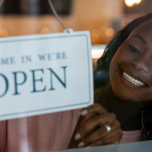 A smiling person flipping a sign to say "Come in we're OPEN" 