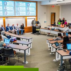 A speaker at a podium in a room of seated others. A digital display behind them with profiles of the panelists.