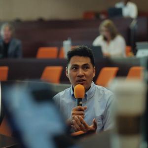 A person seated in an auditorium talking into an orange mic.
