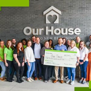 A greoup of people posed in front of the side of a building "Our House" sign above them. Some holding a large check made to "Our House".