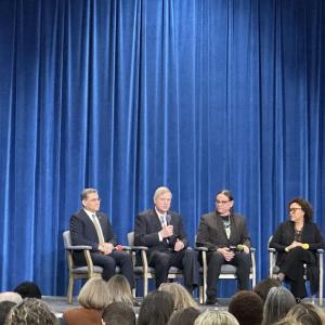 A panel of four seated on a stage in front of a room of people.