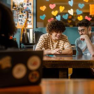 People seated at a table, looking at the same tablet. A person blurred in the foreground using a laptop. Colorful hearts on a window in the background.