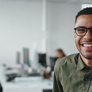 A smiling person looking at the camera, a small group of people at a shared desk behind them, in an office setting