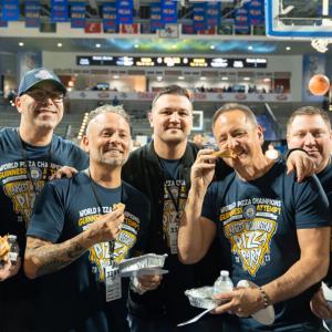 Seven people in matching t-shirts pose for the camera, each has a slice of pizza. The basketball court behind them.
