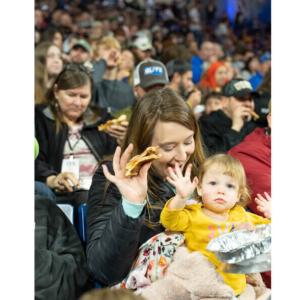 A crowd of people in the stands, close up of an adult and child waving, a piece of pizza in their hands.