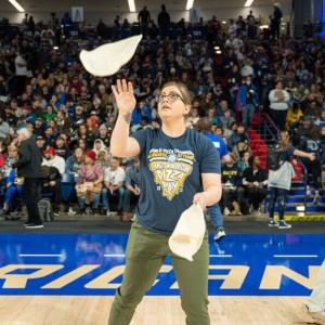 A person standing on a basketball court tossing two pizza dough crusts. A person walking behind them with a microphone. A large crowd in the stands.