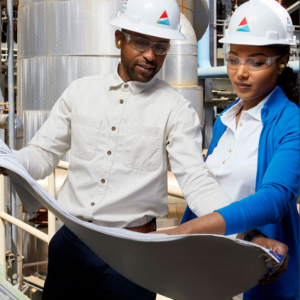 Two people in business casual attire, hard hats and protective eye ware looking over a blueprint. They are standing in a large plant.