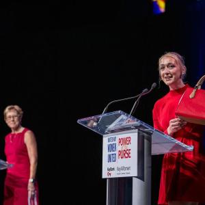 person standing at event podium holding a red purse