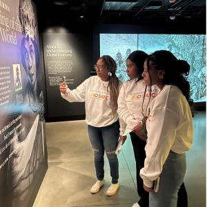 Three women stand in front of museum panels reading history content. One takes a photo with a cell phone as two look on.