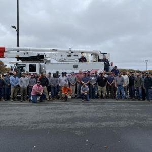 A large group of people posed outside in a parking lot in front of a truck with large extension arm and bucket.
